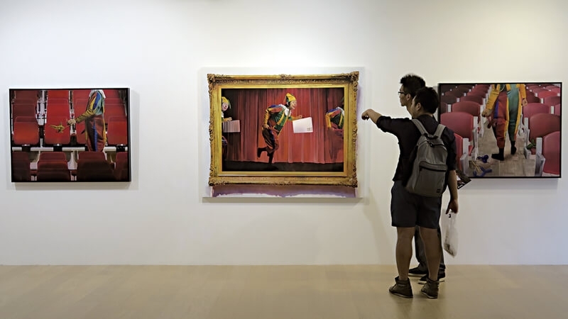 展場一景，觀者駐足於作品前討論。左：《紅幕落下時》．油彩、麻布．130×162 cm．2015、 右：《幻象事實在裝傻》．油彩、麻布．91×116.5 cm．2015。