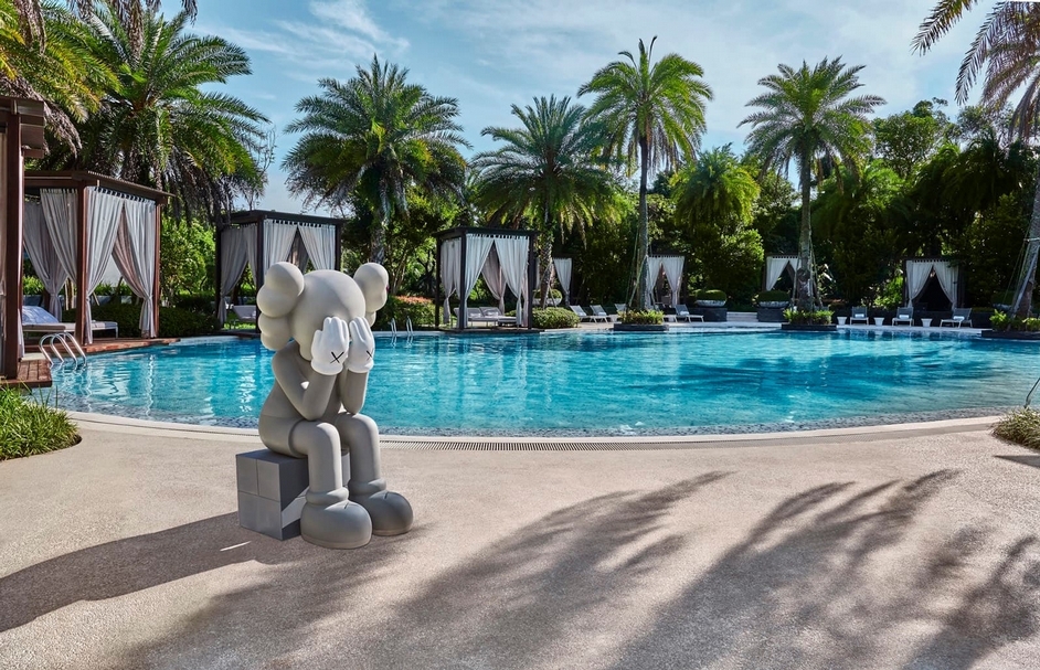 KAWS《同伴（經過）》（灰）來到在熱帶棕櫚樹圍繞的泳池池畔，吸收耀眼陽光的熱情活力。（中誠拍賣提供）
