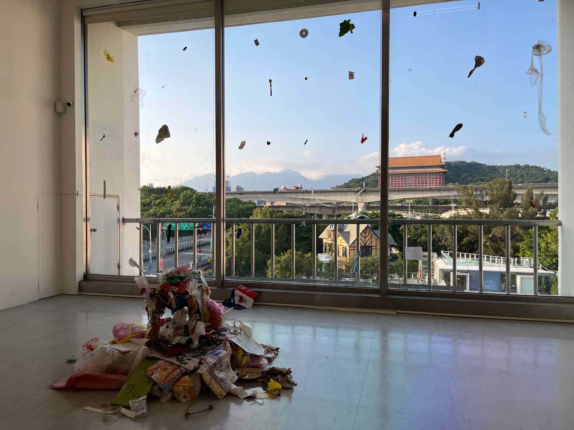 展場中一隻「垃圾狗」與牠扭動一地的「垃圾堆」，搭配背後一大面落地窗上的「垃圾毛毛雨」，一路望向窗外的藍天與圓山。