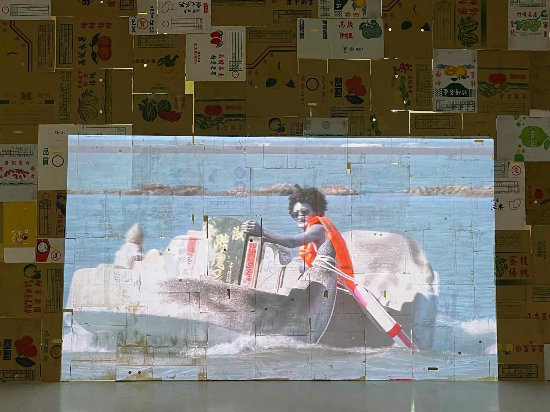 最後的小展場，一大面「攤平的各種水果箱」拼成的投影牆，播映一座因地層下陷，而泡在水裡的墓園。