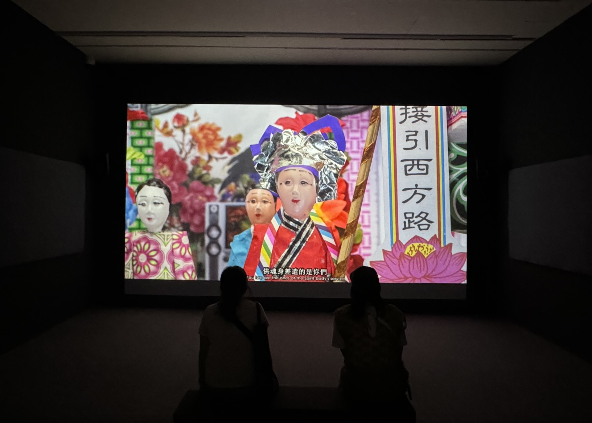 圖為蘇育賢《花山牆》於「黑水」展出現場。作品以紙紮工藝與影像敘事交織，透過「金童玉女樂隊」的引領，將個人命運與國家歷史並置。燃燒的火焰象徵死亡，也隱喻臺灣自明清、日治、國民政府至當代資本主義的殖民軌跡；日式巴洛克山牆則指向歷史遺緒中持續燃燒…