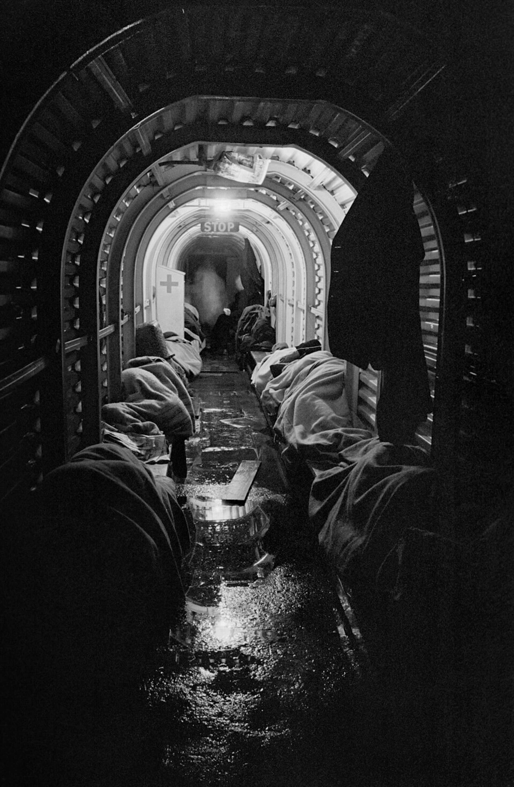 比爾・布蘭特（Bill Brandt）的攝影作品《倫敦防空壕中的避難者》（1940 ）。照片攝於二戰期間德軍空襲（Blitz）下的倫敦，民眾在遭洪水淹沒的地下防空壕中休息，地面以木板臨時鋪設以避開積水，後方可見急救箱。（© Public D…