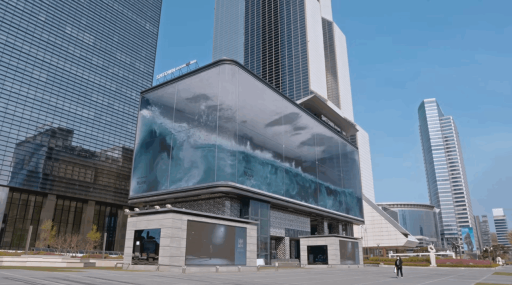 d’strict《Public Media Art #1_WAVE》（2020），首爾 COEX K-Pop 廣場。巨浪以變位錯視在寬約80公尺的曲面LED螢幕上翻湧，成為城市螢幕轉為觀看媒介的代表性場景。（取自d’…